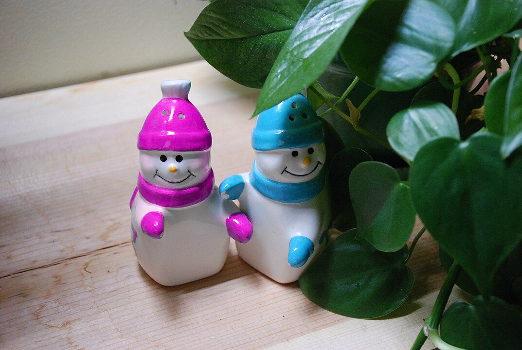 Two ceramic snowman figurines, one with a pink hat and scarf, the other with a blue hat and scarf, standing side by side on a wooden surface