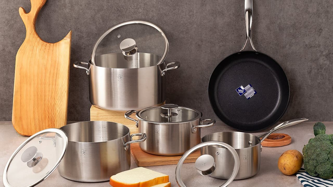 Stainless steel cookware set, includes pots and pans with glass lids, nonstick frying pan hanging on wall, wooden cutting board