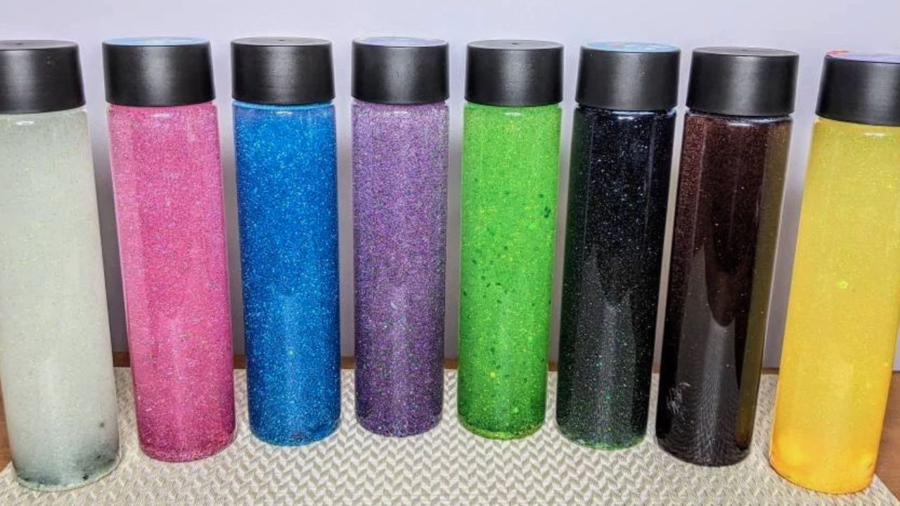 Eight glitter sensory bottles in various colors—white, pink, blue, purple, green, black, brown, and yellow—lined up on a textured surface with black caps and a light background