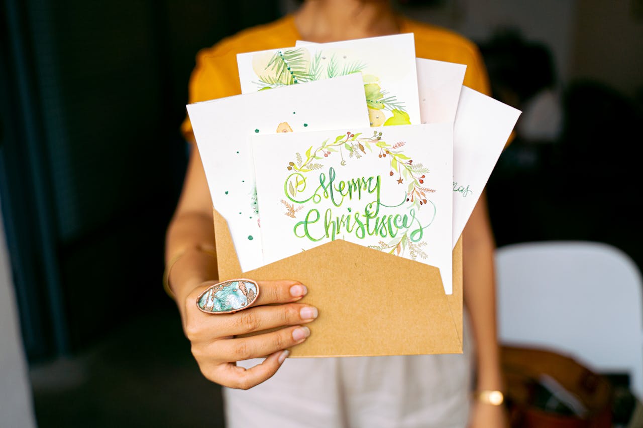 Person holding a brown envelope filled with festive illustrated Christmas cards, one card reads "Merry Christmas