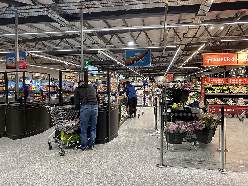 Supermarket checkout area, shoppers with carts in line, bright overhead lighting, visible produce section to the right, high ceilings with hanging signs