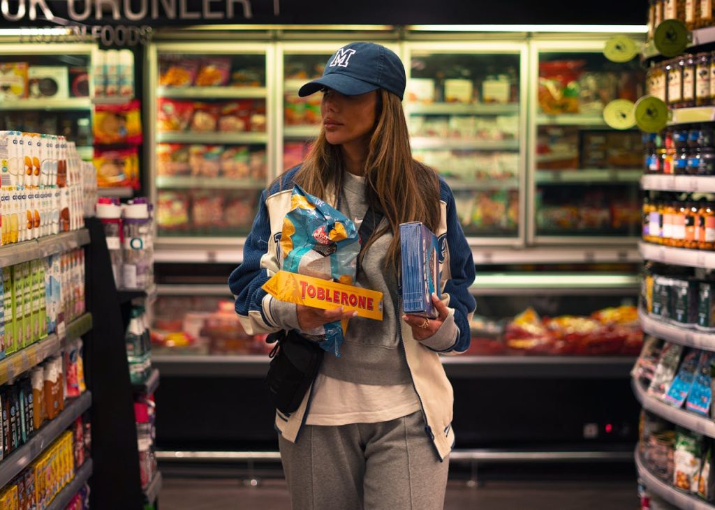 Woman wearing a cap and casual clothes, holding a Toblerone bar and frozen food items, shopping in a dimly lit grocery store aisle, standing between shelves of drinks and snacks, suggesting late-day or evening shopping
