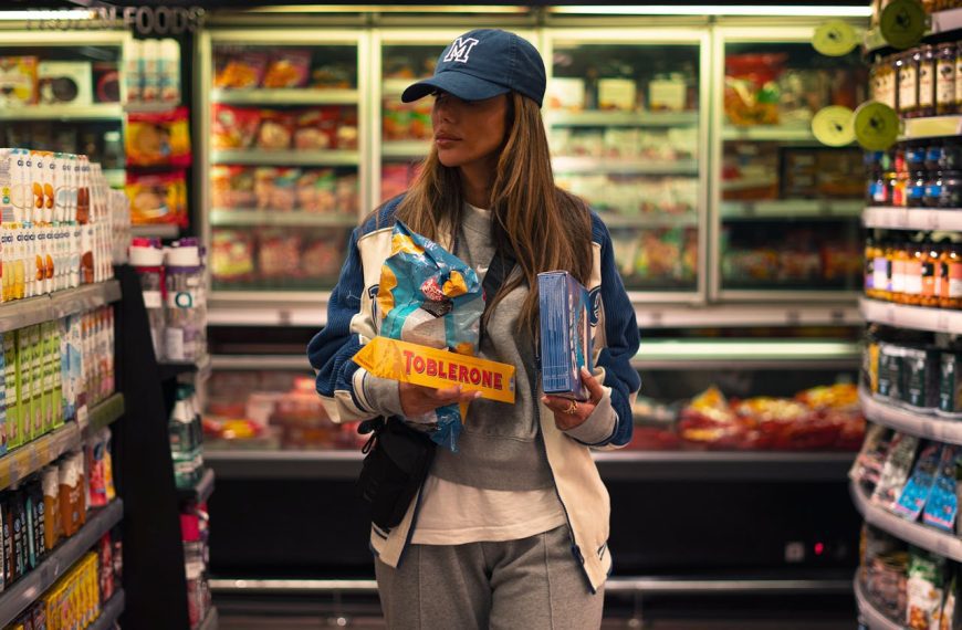 Woman wearing a cap and casual clothes, holding a Toblerone bar and frozen food items, shopping in a dimly lit grocery store aisle, standing between shelves of drinks and snacks, suggesting late-day or evening shopping