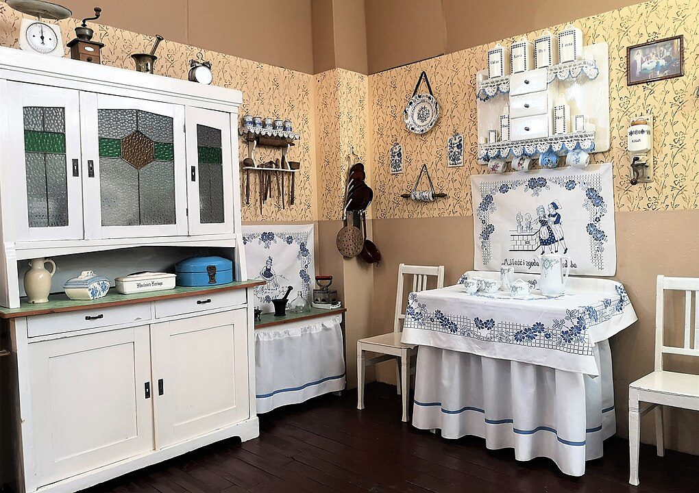 Vintage kitchen scene, white sink with blue floral retro print skirt, white wooden hutch with glass doors, antique dishes and decor, matching white chairs, warm beige walls with framed embroidery, cozy and nostalgic ambiance