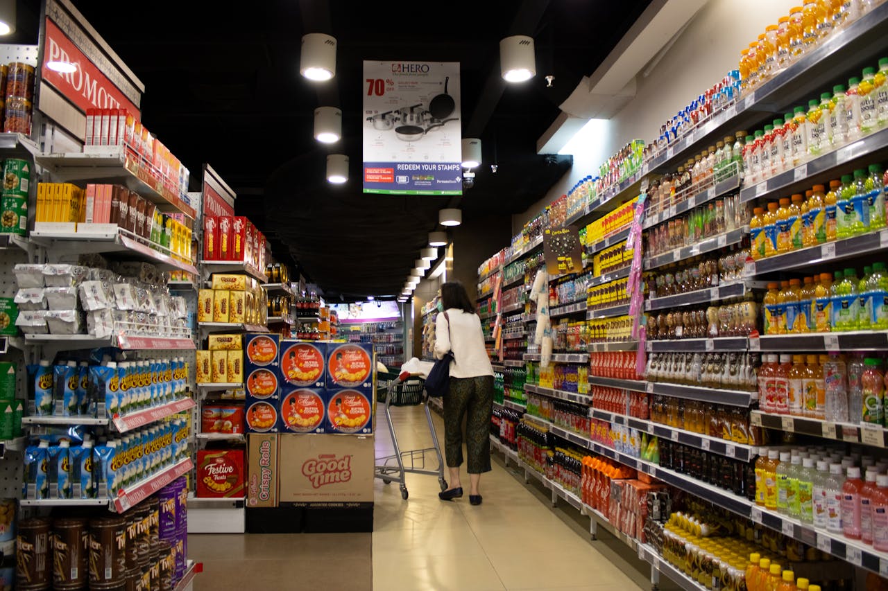 Woman grocery shopping, walking down a brightly lit supermarket aisle, shelves filled with snacks and beverages on both sides, pushing a shopping cart, appearing focused on her path, snacks and drinks prominently displayed around her