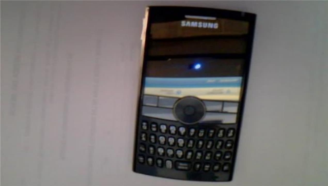 Old Samsung smartphone, candy bar style, full QWERTY keyboard, small screen with navigation button in the center, photographed on a white surface under dim lighting