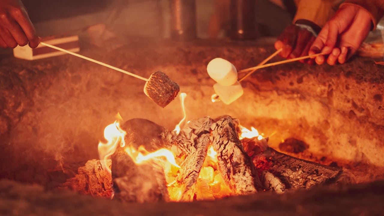 Close-up of people roasting marshmallows over a glowing campfire with charred logs and visible flames