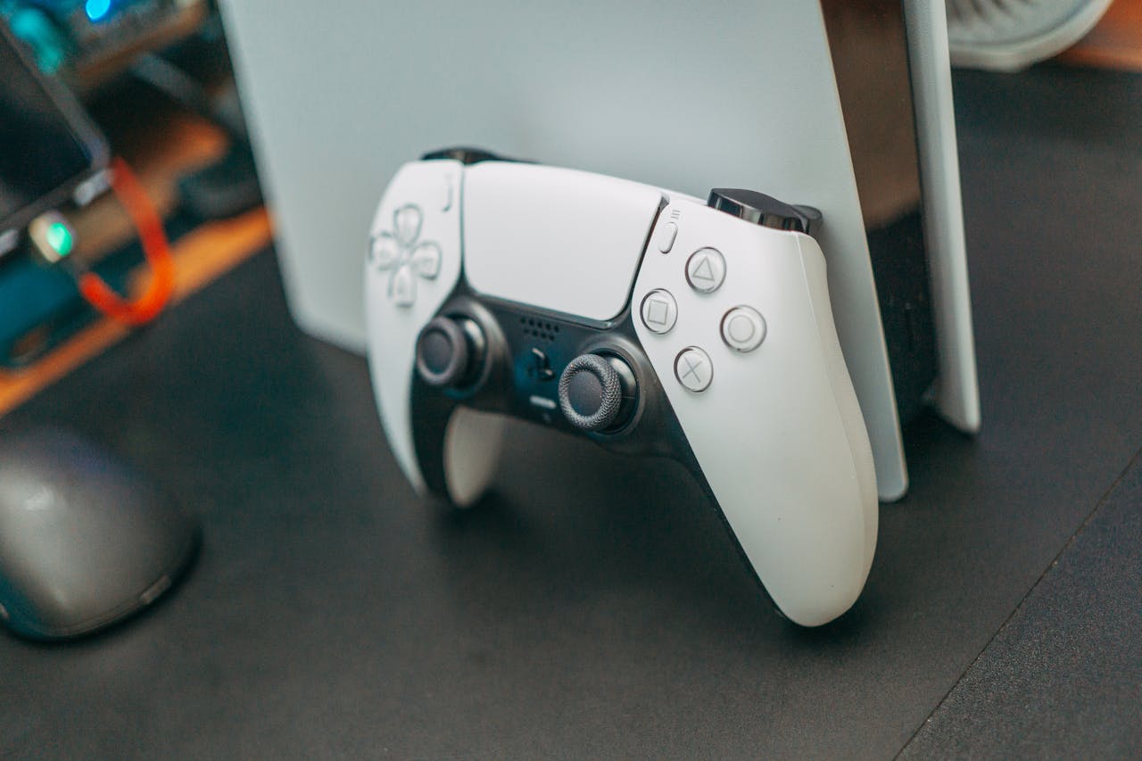 White PlayStation 5 DualSense controller resting against a PlayStation 5 console, on a dark gaming desk with accessories in the background