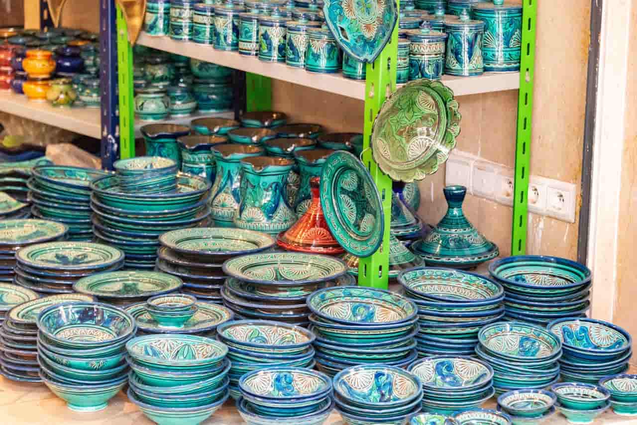 Stacks of traditional Moroccan ceramic plates and bowls, predominantly in turquoise and blue, with intricate white and green patterns, displayed on shelves in a pottery shop