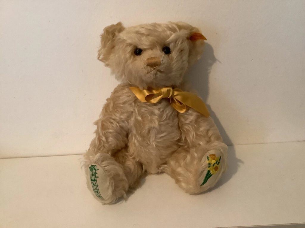 Beige teddy bear, seated upright, yellow ribbon tied around neck, green embroidered details on feet, soft textured fur, plain white background