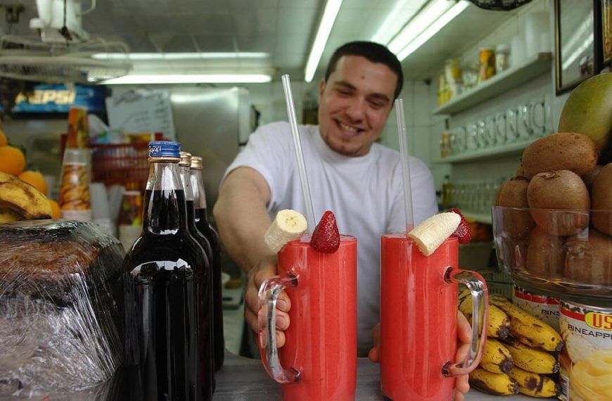 A smiling worker in a white shirt holds two tall glasses of bright red smoothies garnished with banana slices and strawberries. Dark glass bottles and fresh fruit surround him in what appears to be a juice shop
