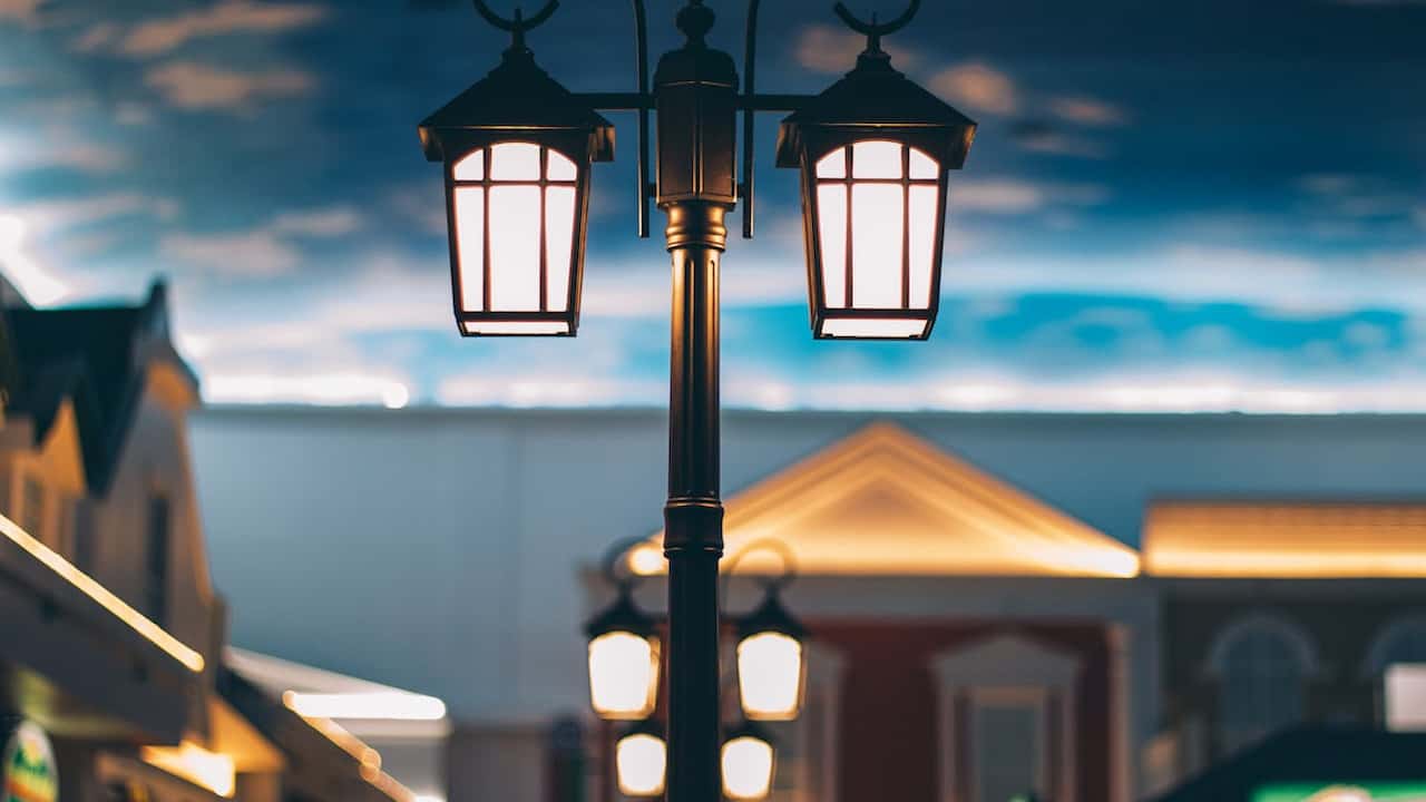 Ornate three-headed streetlamp glowing warmly in a shopping area with blurred storefronts and a painted sky ceiling in the background