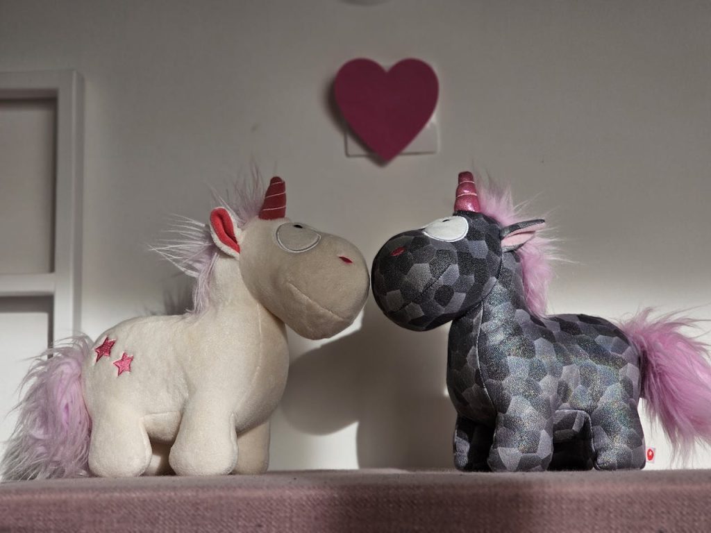 Two stuffed unicorn toys facing each other, one white with red stars and the other dark patterned, both with pink manes and tails, red and pink horns