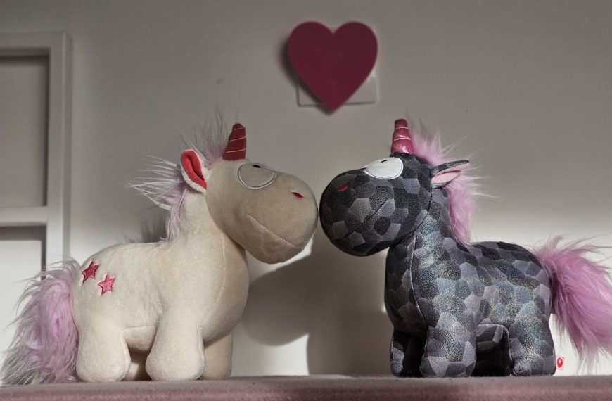 Two stuffed unicorn toys facing each other, one white with red stars and the other dark patterned, both with pink manes and tails, red and pink horns
