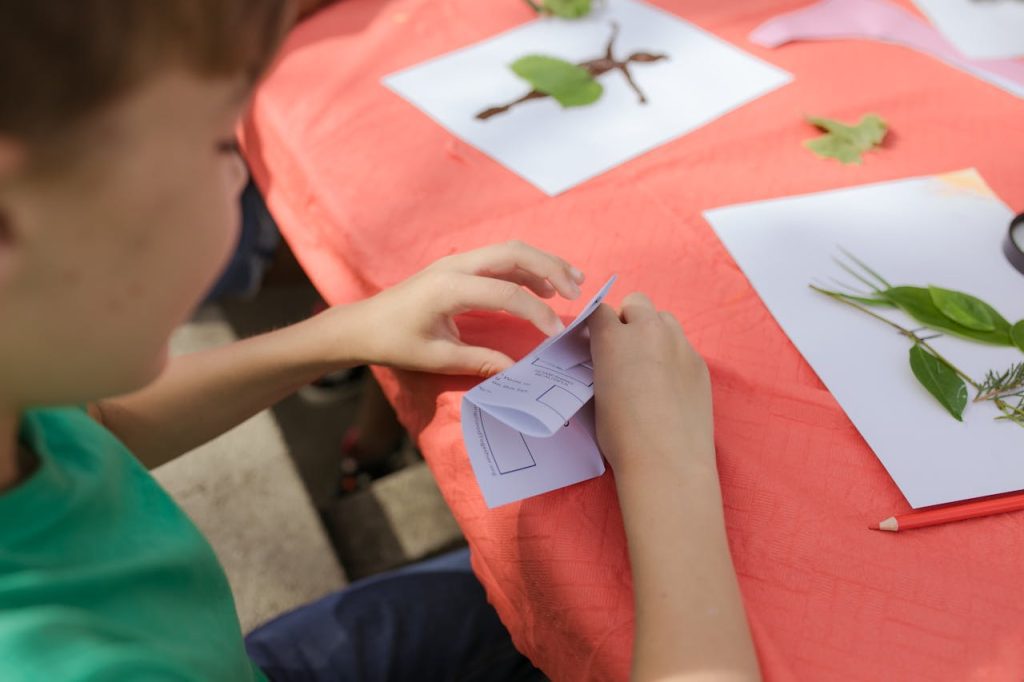 Child working on a craft project, folding paper with printed shapes, surrounded by white papers with arranged leaves and natural materials, outdoor setting