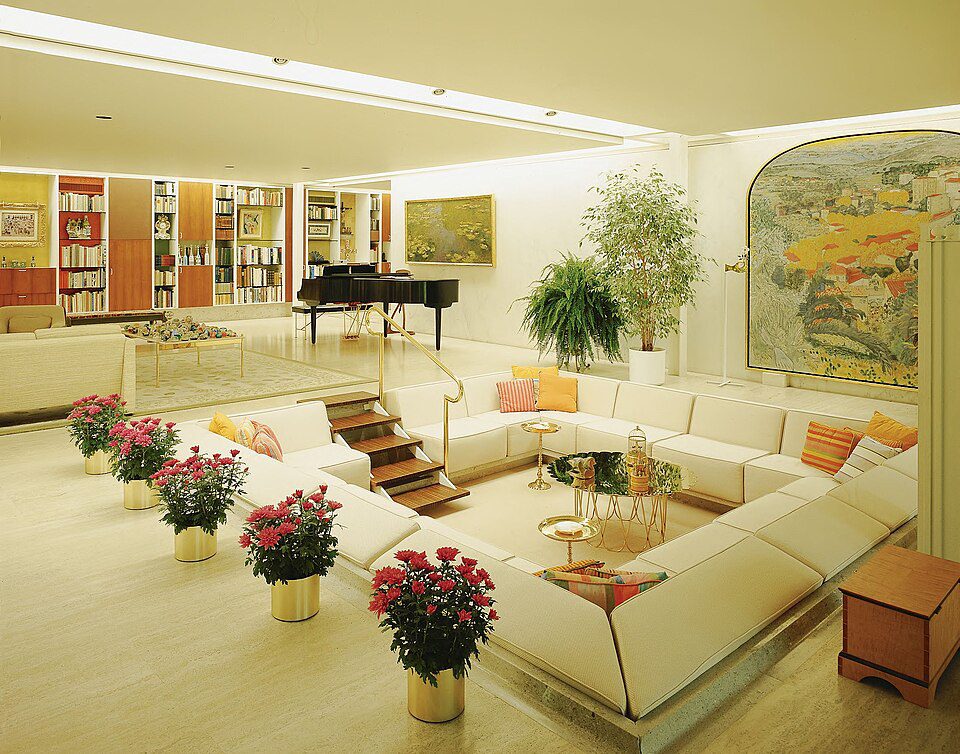 Sunken white sectional sofa with orange and beige cushions, potted pink flowers around the seating area, large indoor plants, colorful abstract art on the walls, grand piano in the background, soft ambient lighting