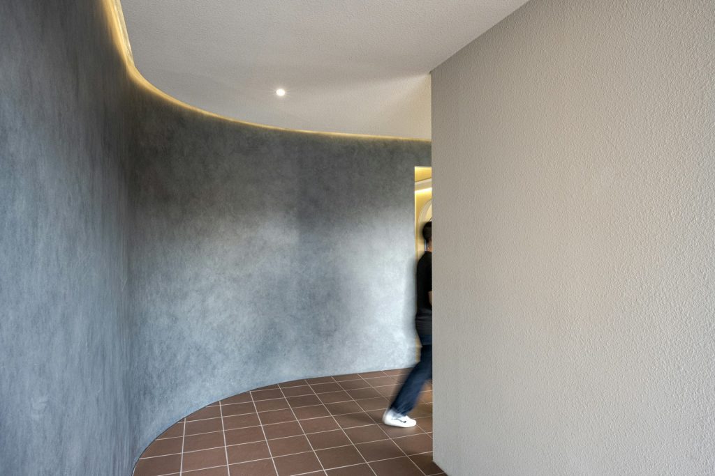 Curved hallway interior, textured gray wall, smooth beige wall, overhead recessed lighting, brown tiled floor, person partially visible walking, modern minimalist architecture
