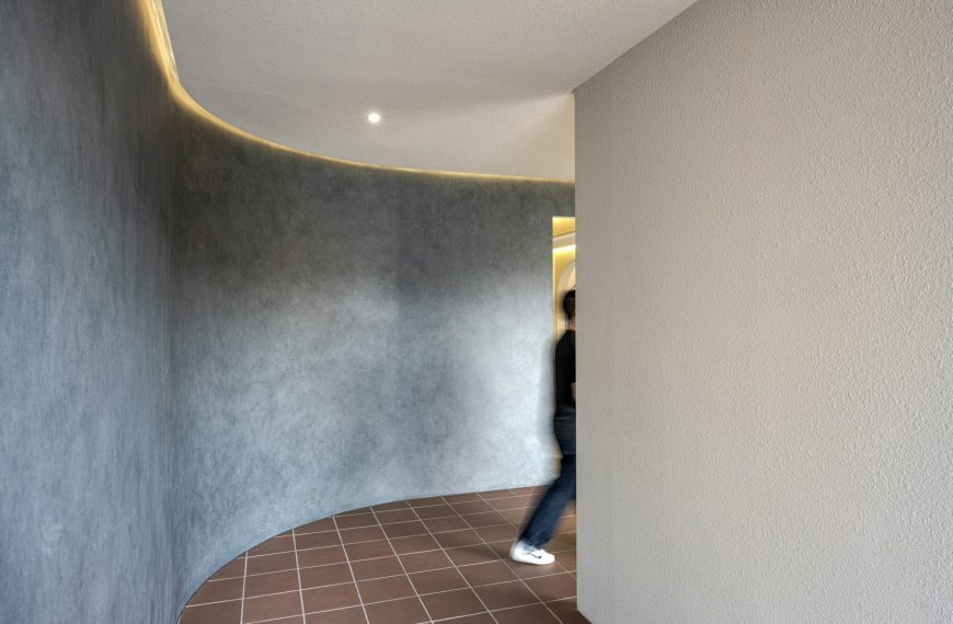 Curved hallway interior, textured gray wall, smooth beige wall, overhead recessed lighting, brown tiled floor, person partially visible walking, modern minimalist architecture