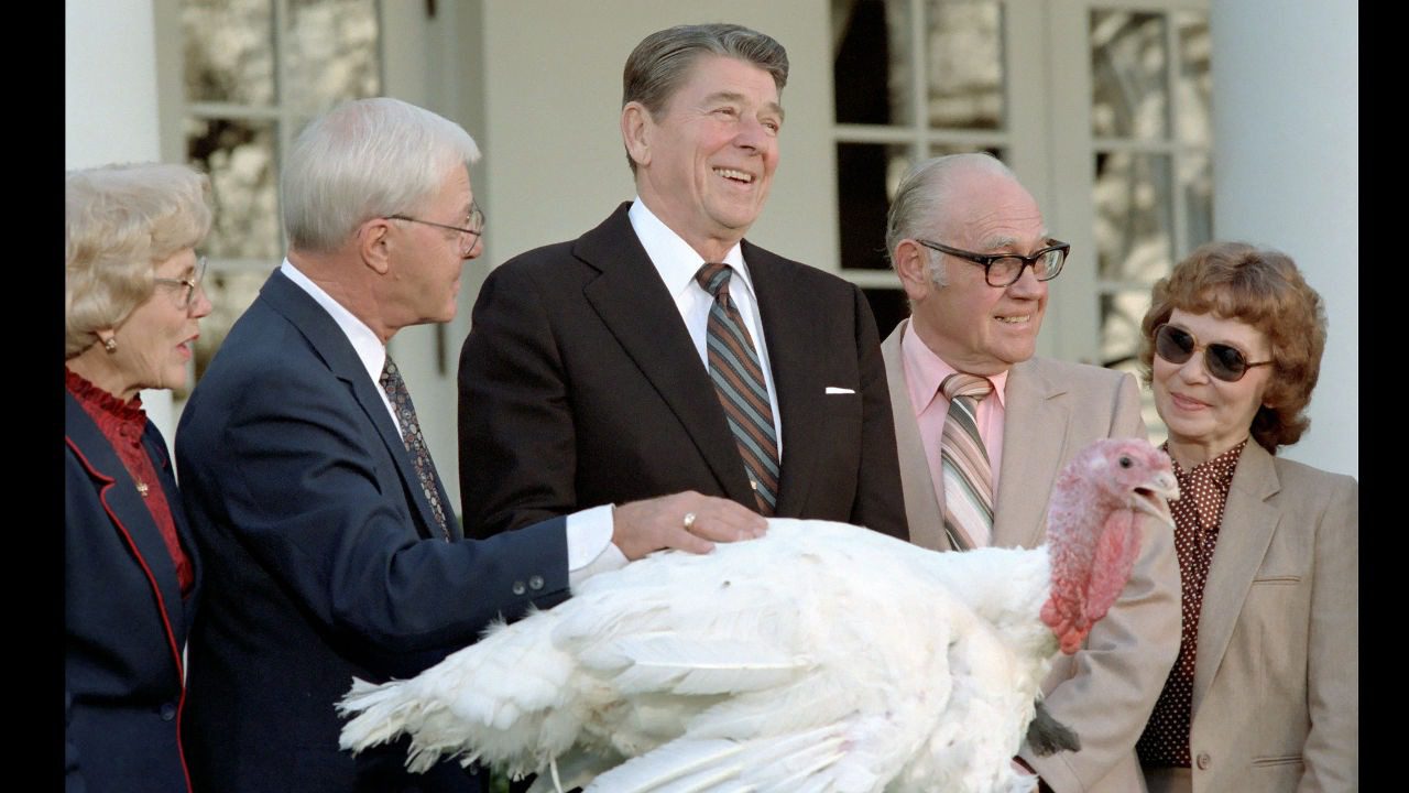 Group of people, including a suited man smiling, standing beside a large white turkey in an outdoor ceremony