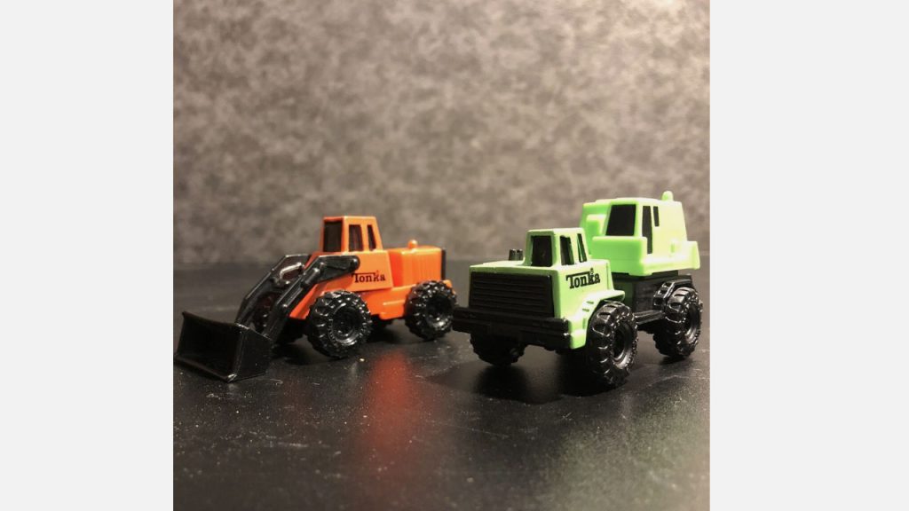 Two small Tonka toy trucks on a black surface, one green flatbed and one orange loader with black scoop