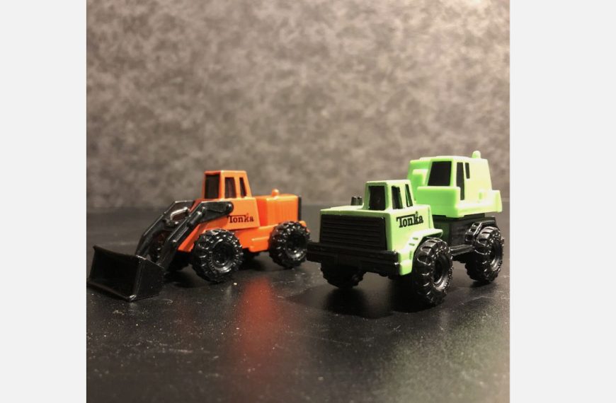 Two small Tonka toy trucks on a black surface, one green flatbed and one orange loader with black scoop