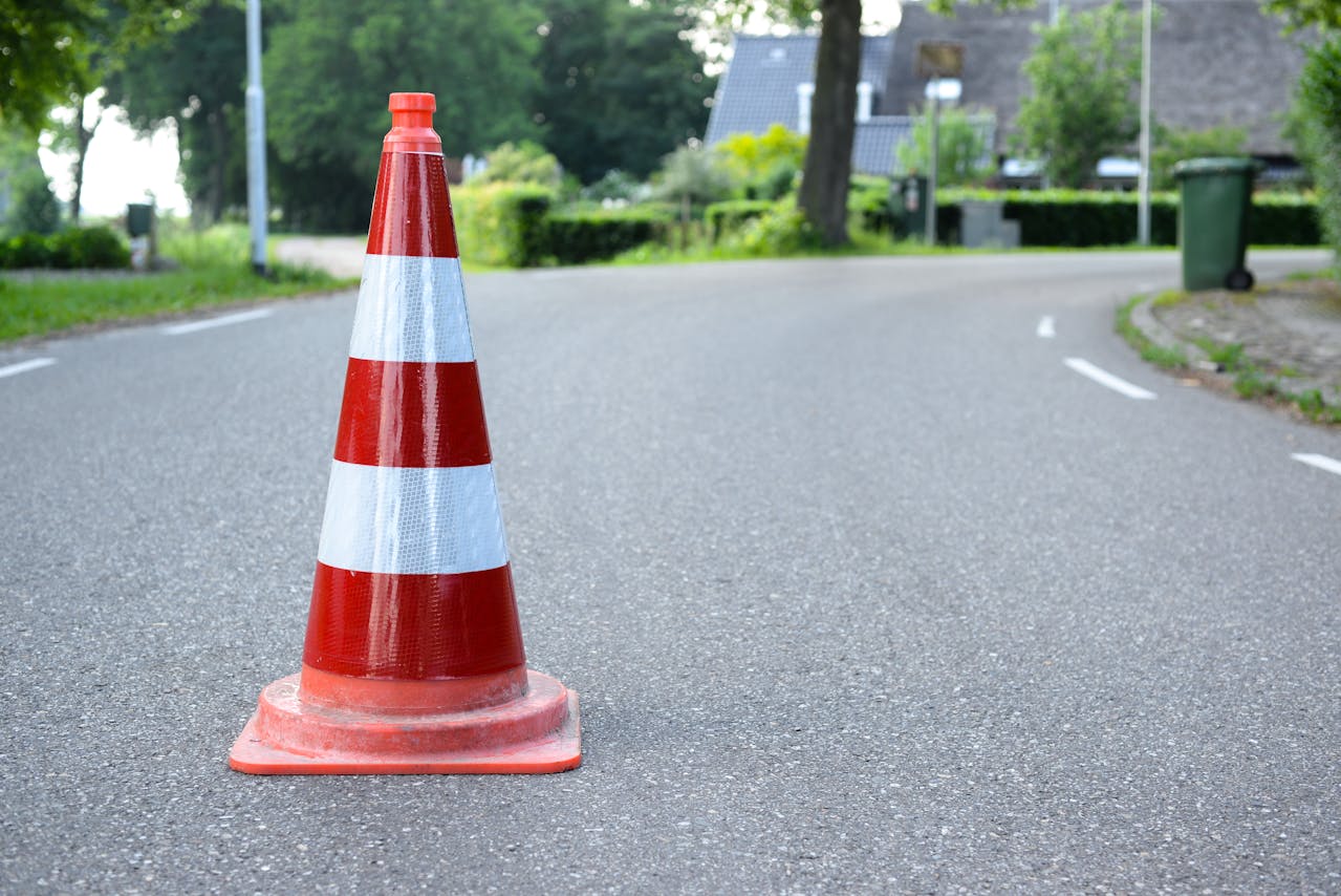 Red and white striped traffic cone placed on an asphalt road in a residential neighborhood with trees and houses in the background.