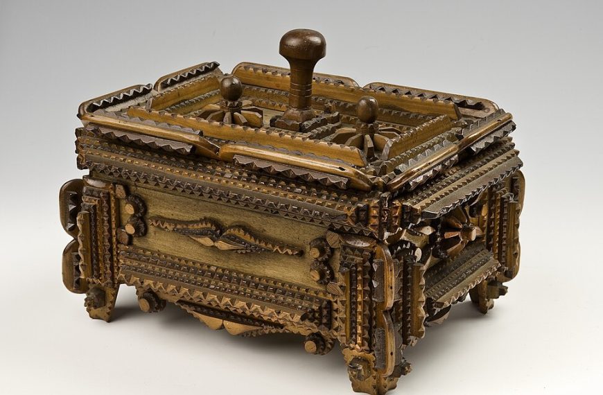 Ornate wooden box with intricate carvings, rectangular shape, decorative finial on lid, elevated base, antique finish, placed against a plain light background