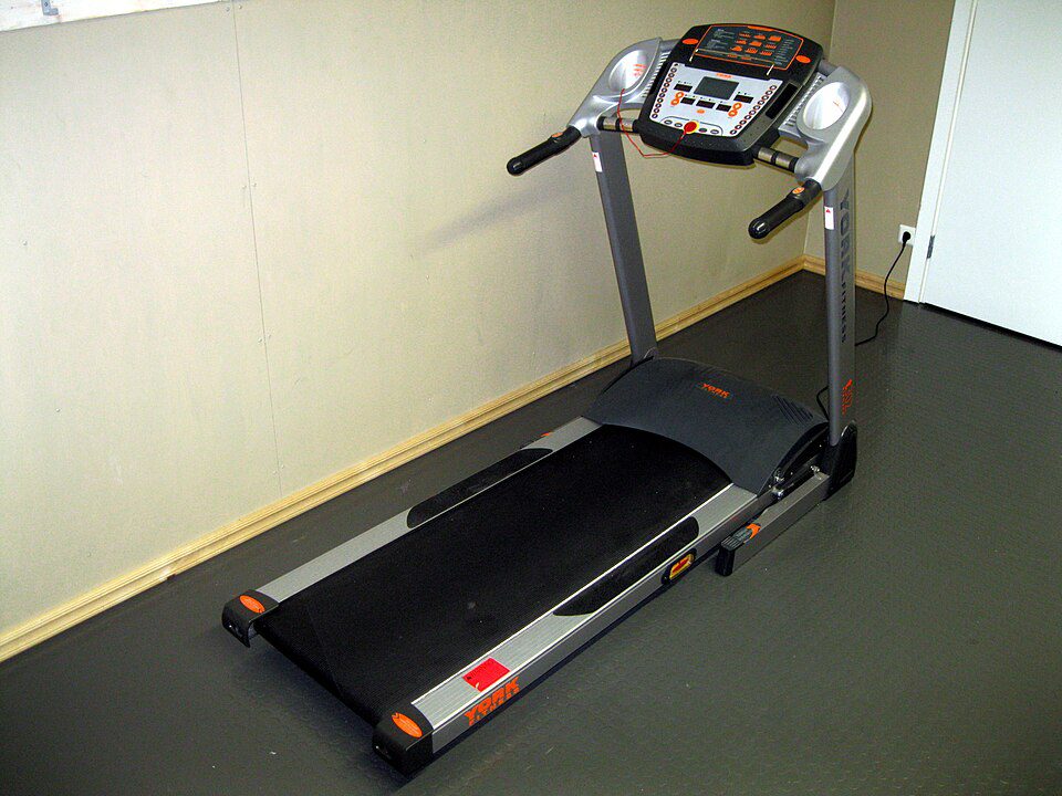 Treadmill positioned in a corner of a room, digital control panel with buttons and display screen, handrails on both sides, black running belt with red accents, yellow trim along the floor edge, suitable for home indoor workouts