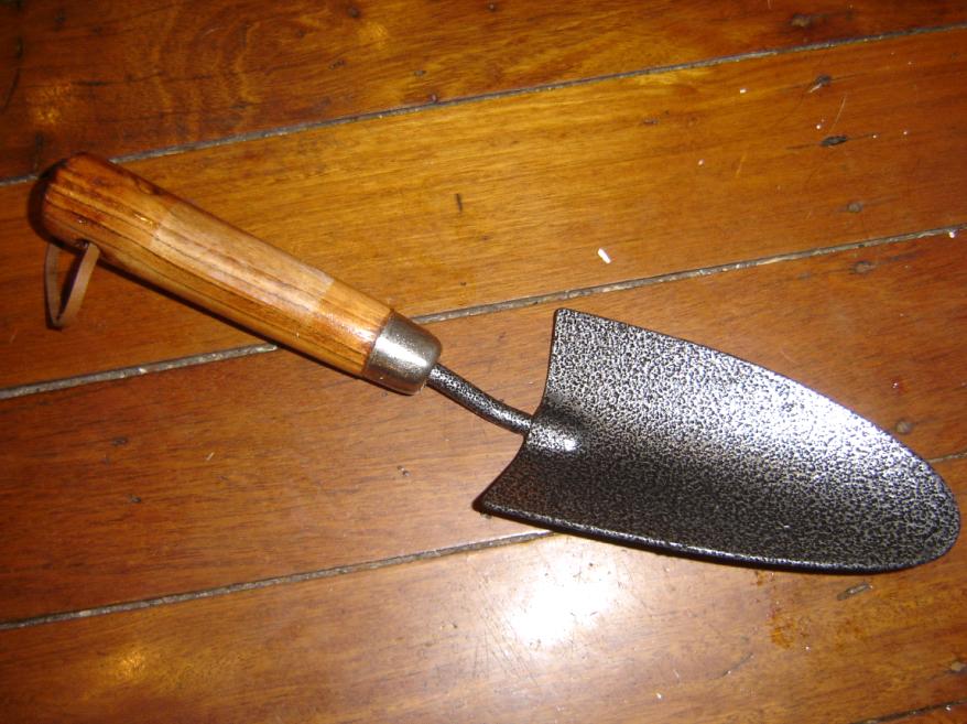 Hand trowel with a wooden handle, black metal scoop-shaped blade, placed on a wooden surface, used for gardening or digging small holes