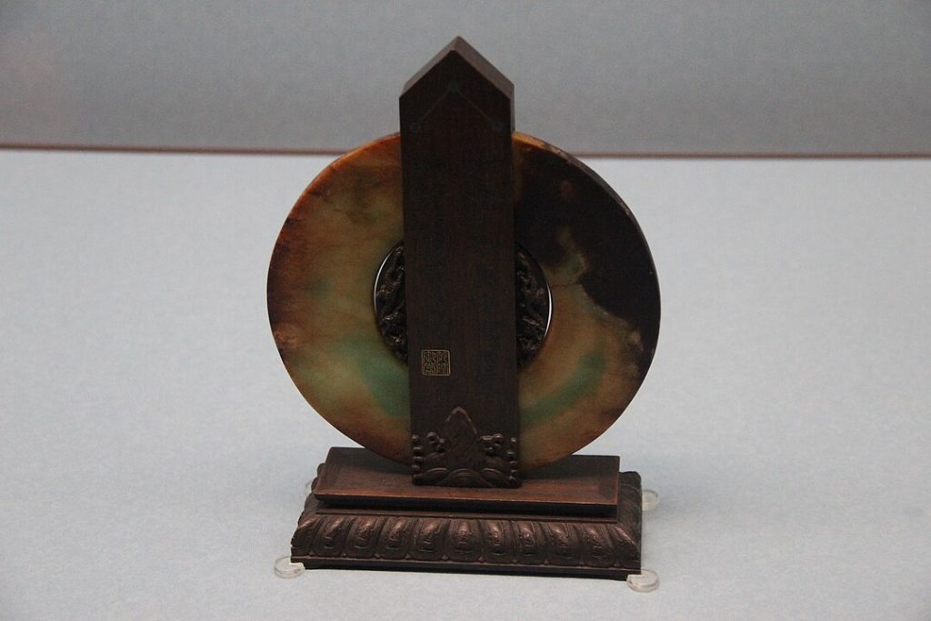 An antique round jade disc with uneven coloration, mounted vertically on an ornate dark wooden stand, photographed against a neutral gray background