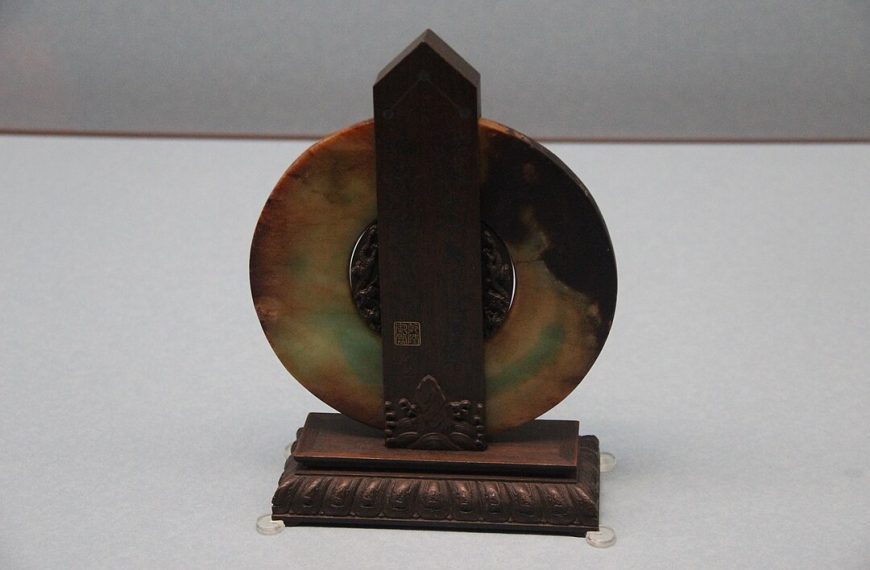 An antique round jade disc with uneven coloration, mounted vertically on an ornate dark wooden stand, photographed against a neutral gray background