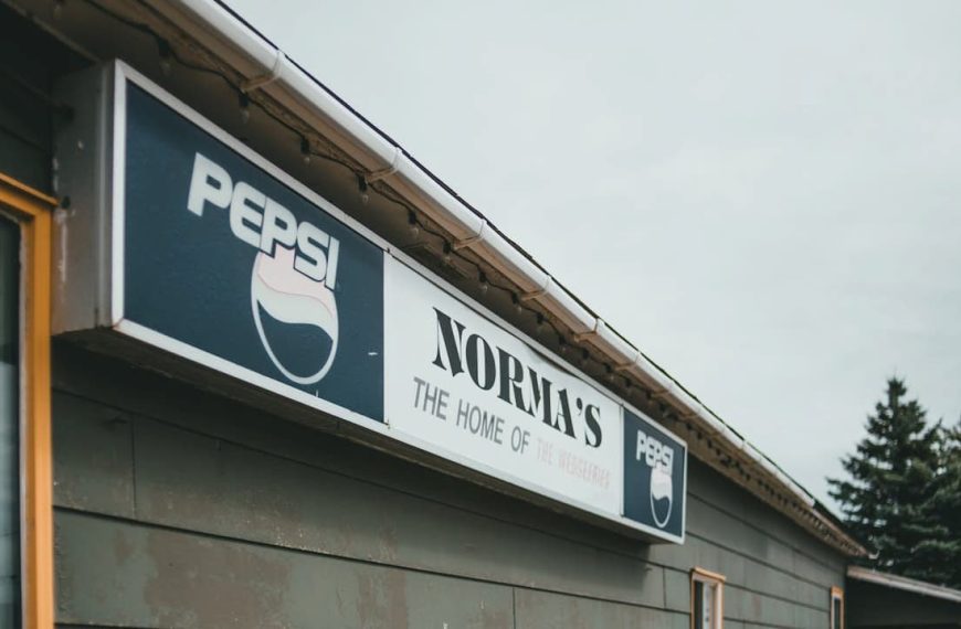 Close-up of a vintage storefront sign showing "PEPSI" logos and the name "NORMA'S THE HOME OF THE WEDGEBURGER" on a weathered building exterior