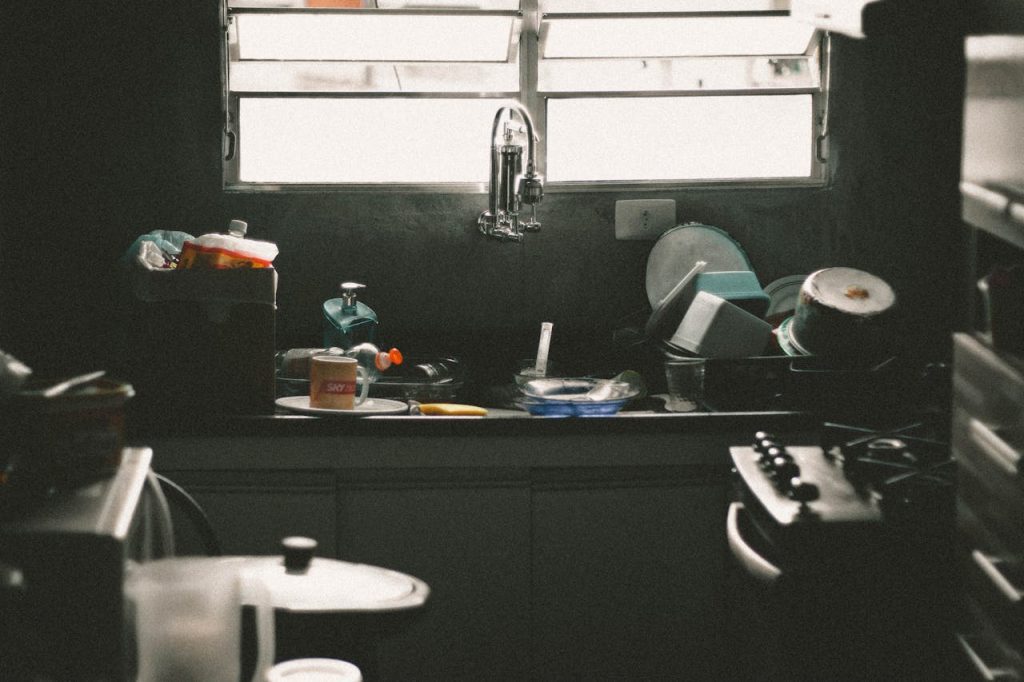 Sink full of unwashed dishes, plastic containers, and utensils under a window with horizontal blinds