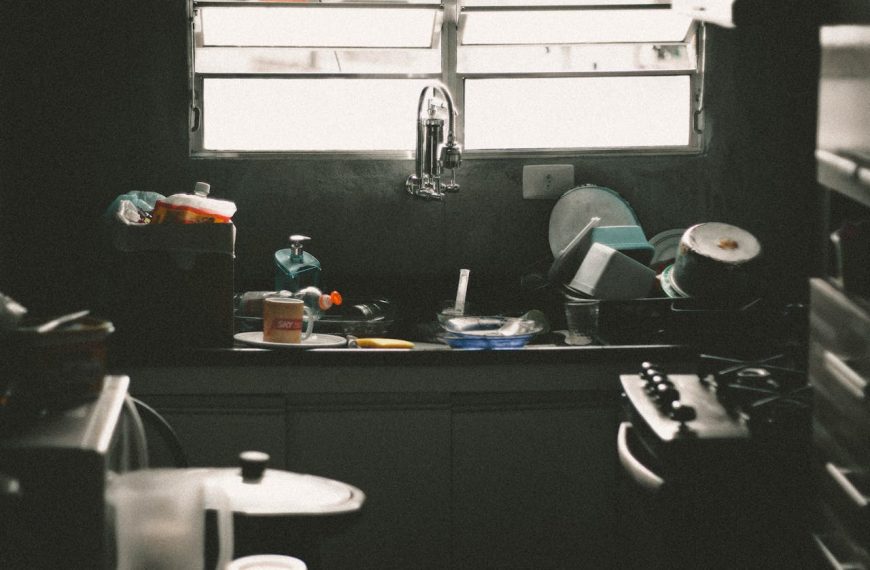 Sink full of unwashed dishes, plastic containers, and utensils under a window with horizontal blinds