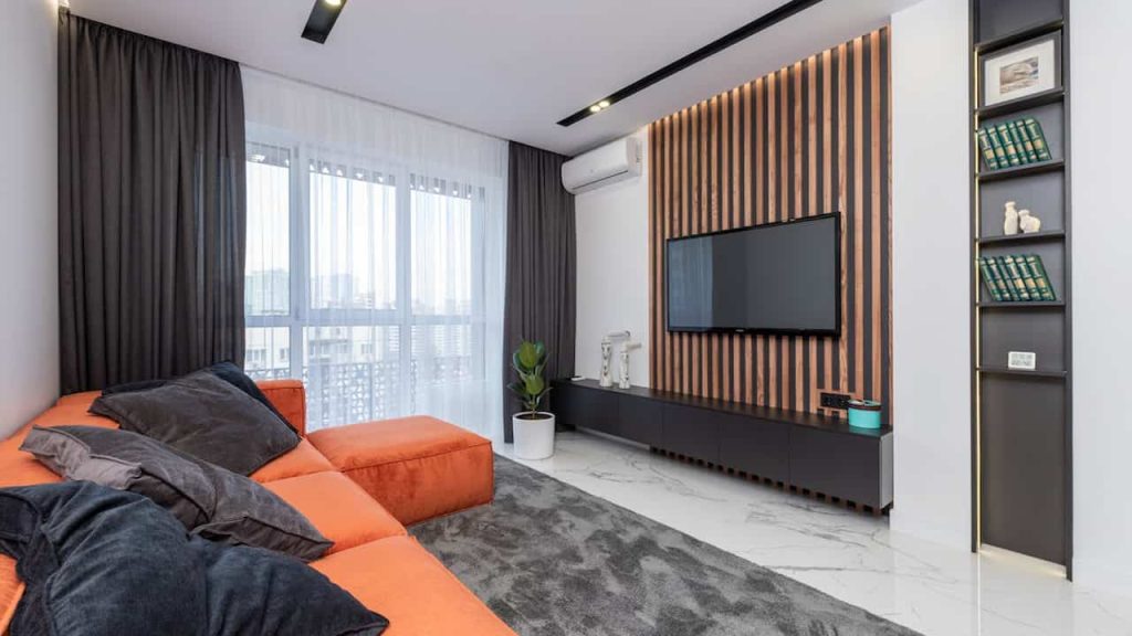 A modern living room with a large orange sectional sofa, a wall-mounted TV against a wood-slat accent wall, a sleek black entertainment unit, and a tall, narrow vertical shelf built into the wall displaying books and décor