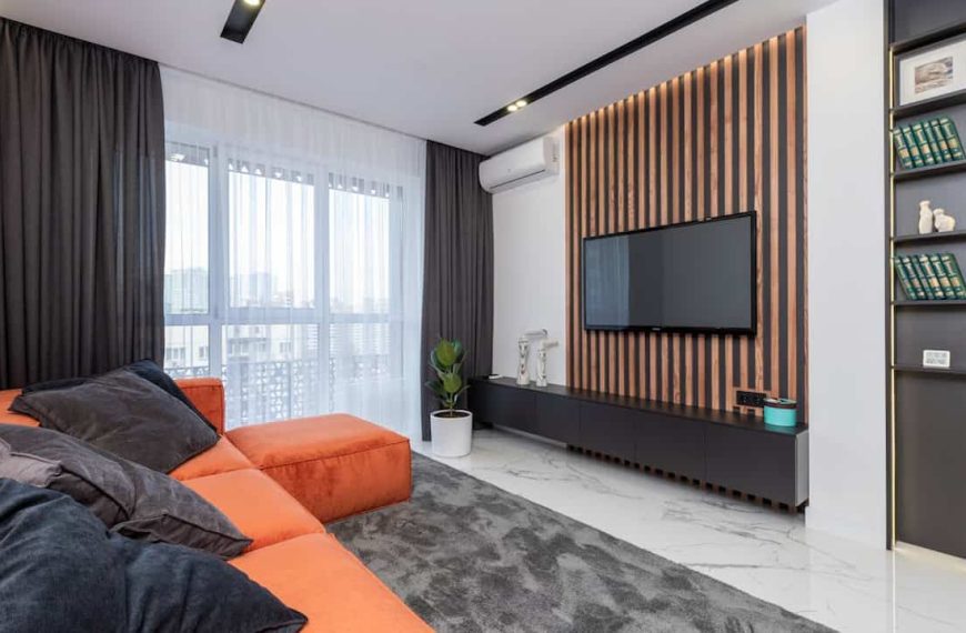 A modern living room with a large orange sectional sofa, a wall-mounted TV against a wood-slat accent wall, a sleek black entertainment unit, and a tall, narrow vertical shelf built into the wall displaying books and décor