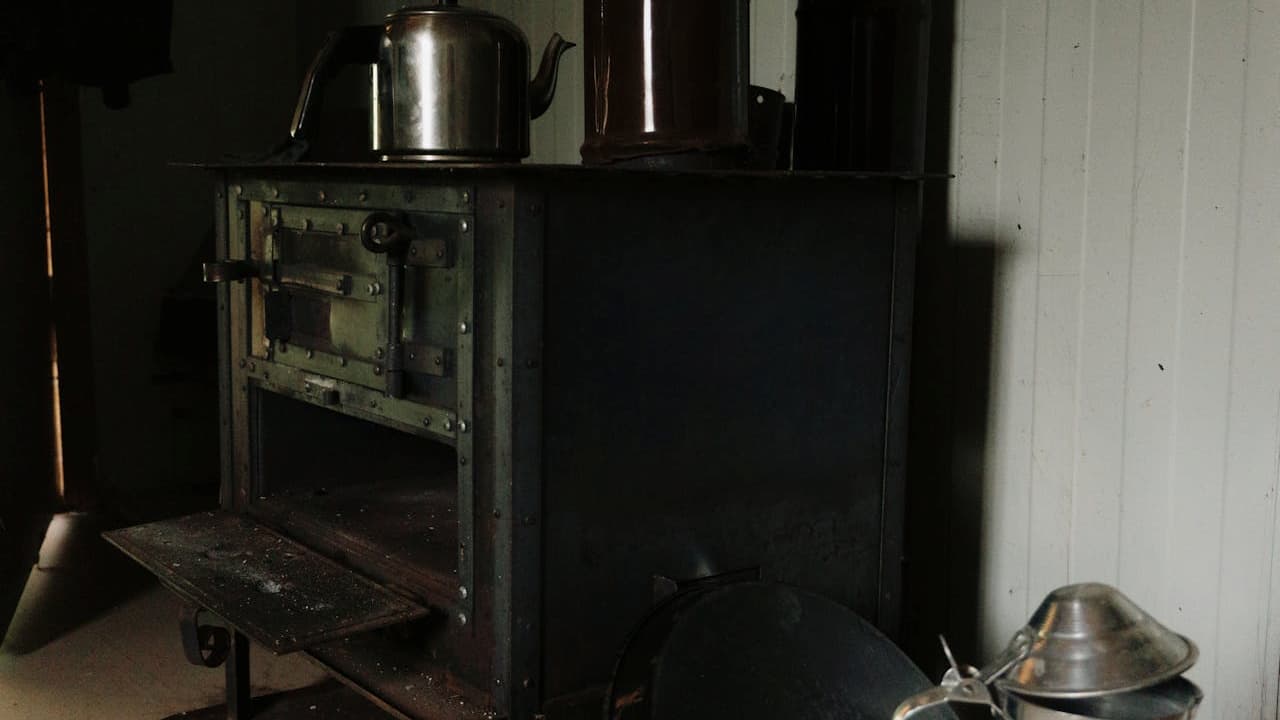 Old-fashioned cast iron wood-burning stove, metal kettle and cylinder on top, open firebox door with ash remnants, dimly lit room with wood-paneled walls, vintage cooking setup, rustic and historical atmosphere