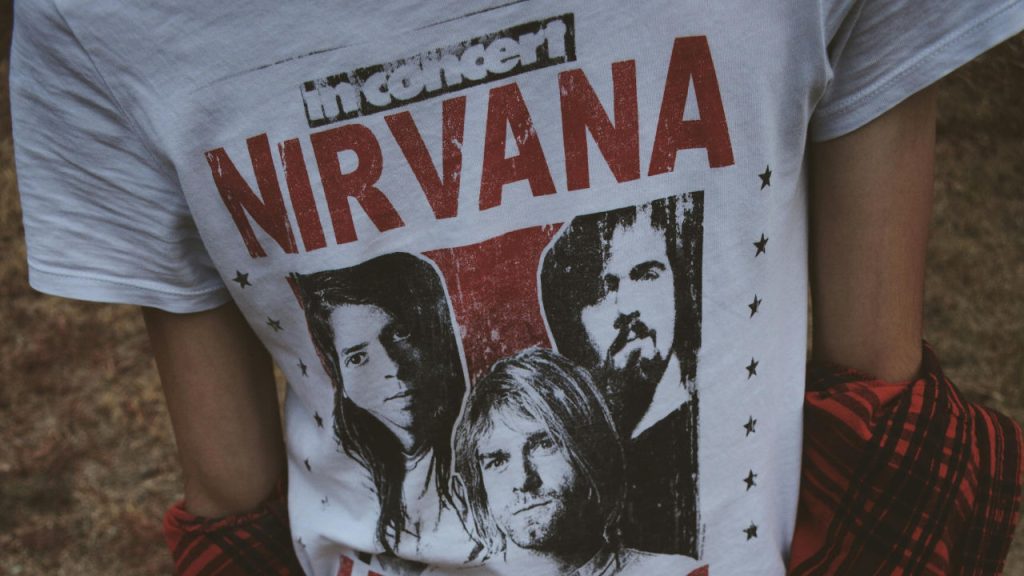 White t-shirt featuring a vintage-style Nirvana band print with distressed red and black design, worn over a red flannel shirt