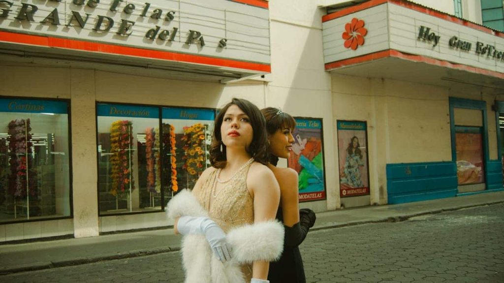 Two women in vintage-style evening gowns, one in a gold dress with a white fur stole and gloves, the other in a black dress with a tiara, standing on a quiet city street in front of a retro storefront