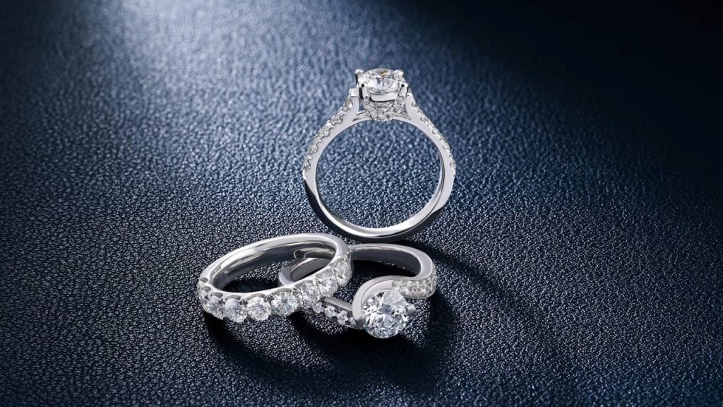 Three elegant diamond rings, silver bands with various stone settings, placed on a dark textured surface, spotlight highlighting their sparkle and clarity
