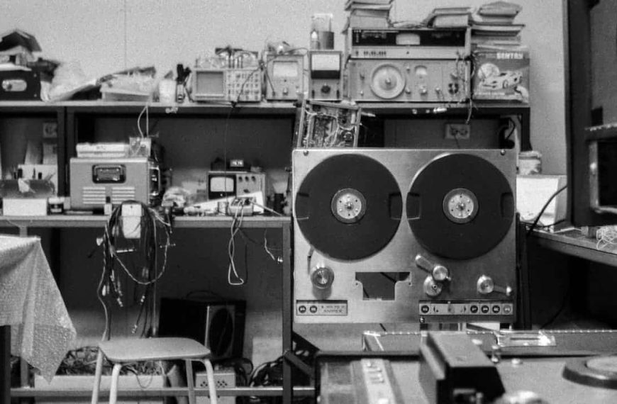 Black and white image of a cluttered electronics workspace, vintage reel-to-reel tape recorder in the foreground, shelves with oscilloscopes, radios, wires, and stacked equipment in the background