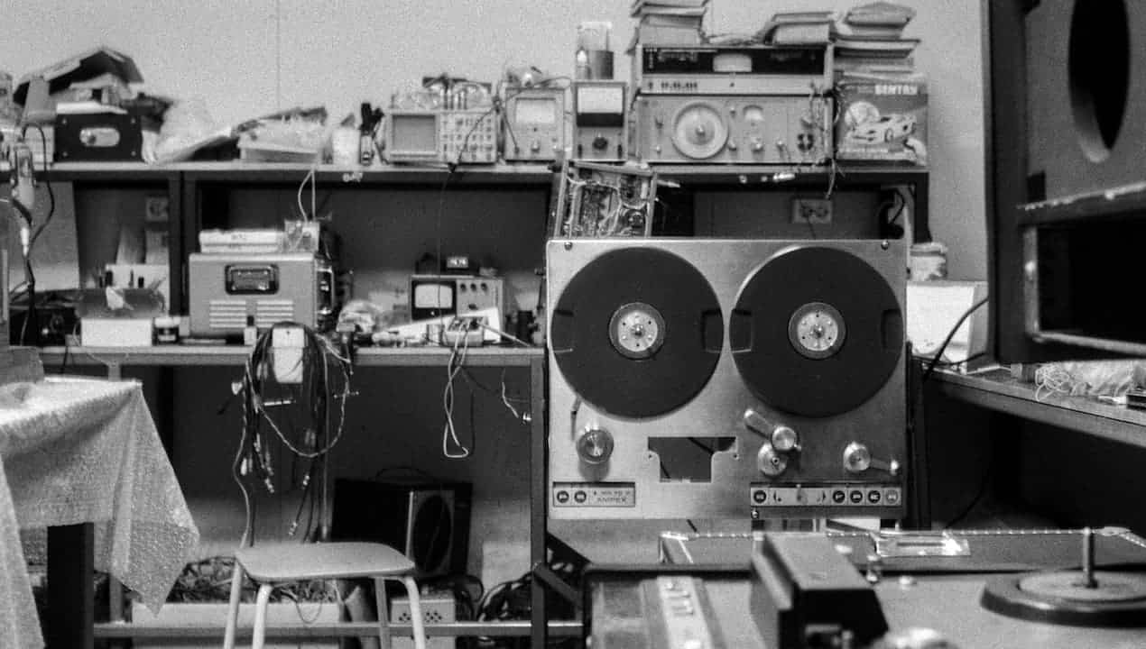 Black and white image of a cluttered electronics workspace, vintage reel-to-reel tape recorder in the foreground, shelves with oscilloscopes, radios, wires, and stacked equipment in the background