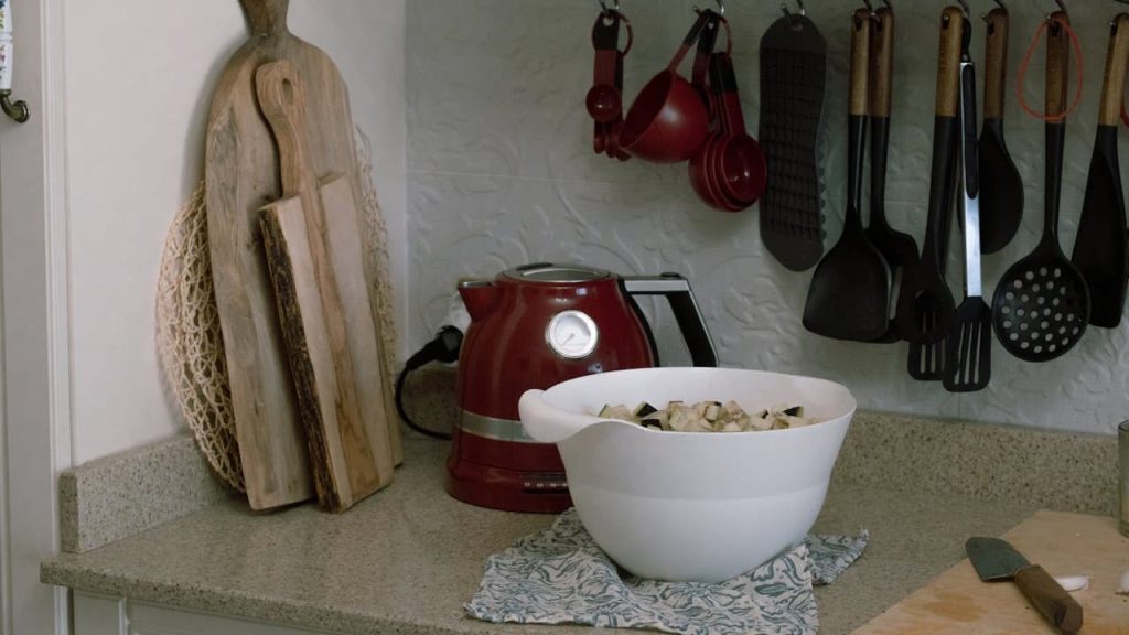 Kitchen countertop with red electric kettle, white mixing bowl filled with chopped vegetables, wooden cutting boards leaning against the wall, hanging kitchen utensils including spatulas and ladles, folded cloth under the bowl, knife on a wooden cutting board.