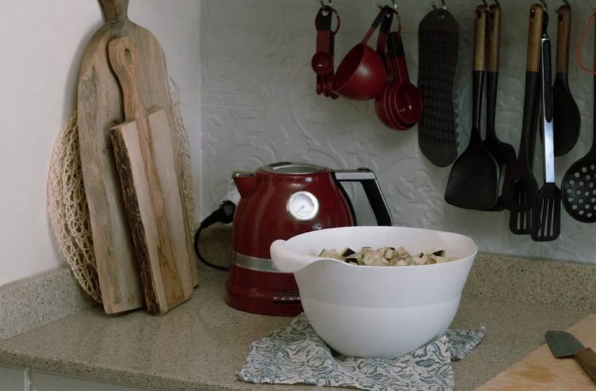 Kitchen countertop with red electric kettle, white mixing bowl filled with chopped vegetables, wooden cutting boards leaning against the wall, hanging kitchen utensils including spatulas and ladles, folded cloth under the bowl, knife on a wooden cutting board.