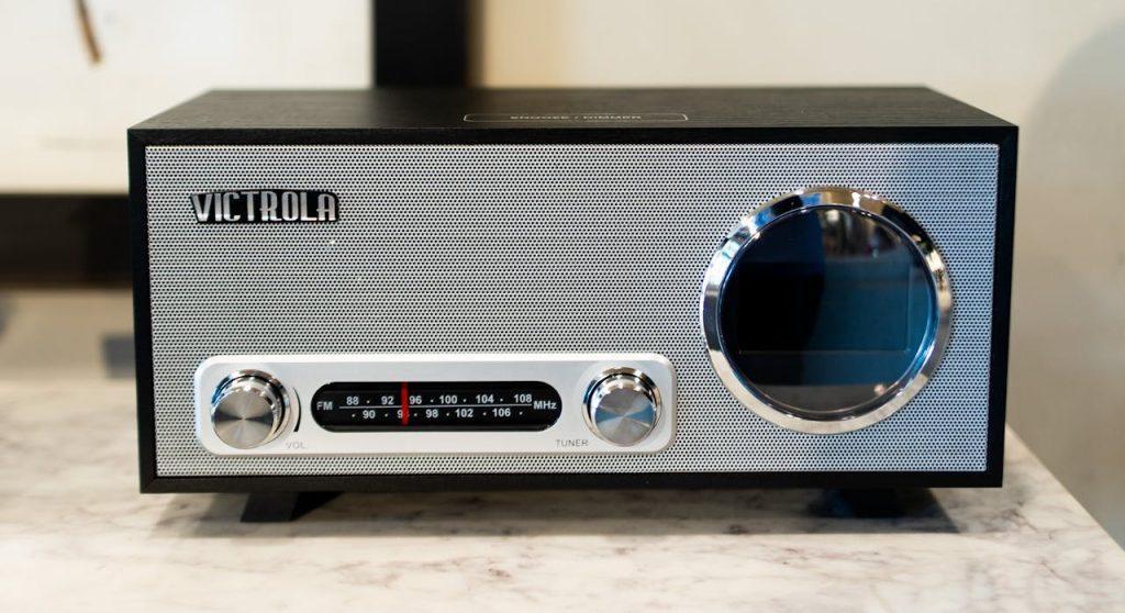 Black and silver Victrola retro radio, front-facing view, two control knobs labeled volume and tuner, FM/AM frequency display in the center, circular speaker on the right, placed on a marble surface indoors