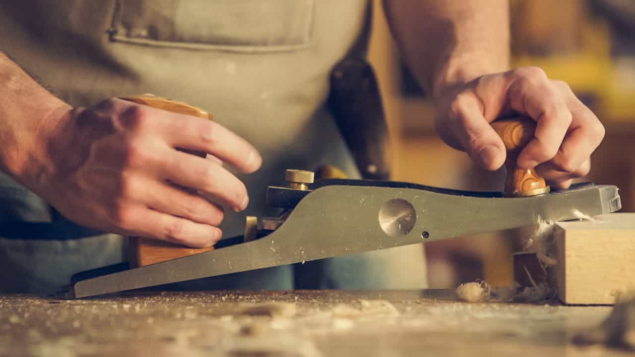 Close-up of hands using a vintage jointer plane, Record No. 7, shaving a piece of wood, woodworking bench, soft warm lighting, apron in background