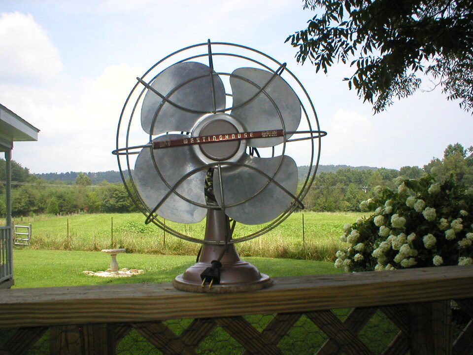 Vintage metal table fan, four-blade design, protective wire cage, mounted on a wooden railing, scenic countryside background, open grassy field, trees and blue sky, peaceful outdoor setting