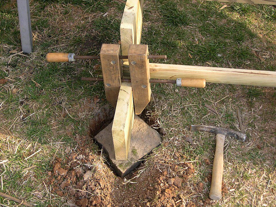 Wooden post held in place by a clamp-style jig, positioned in a dug hole, surrounded by soil and grass