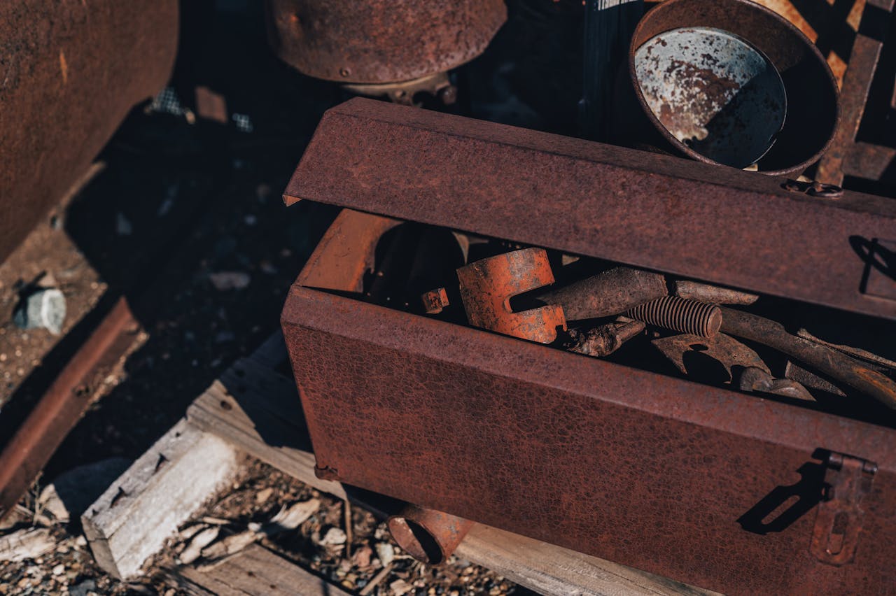 Rusty metal toolbox partially open, filled with old, weathered tools, springs, and metal parts, resting on rough wooden planks