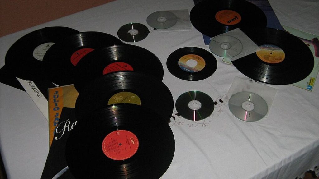 Vinyl records and compact discs scattered on a white surface, multiple black records with colored labels, CDs reflecting light, casual display of music media collection